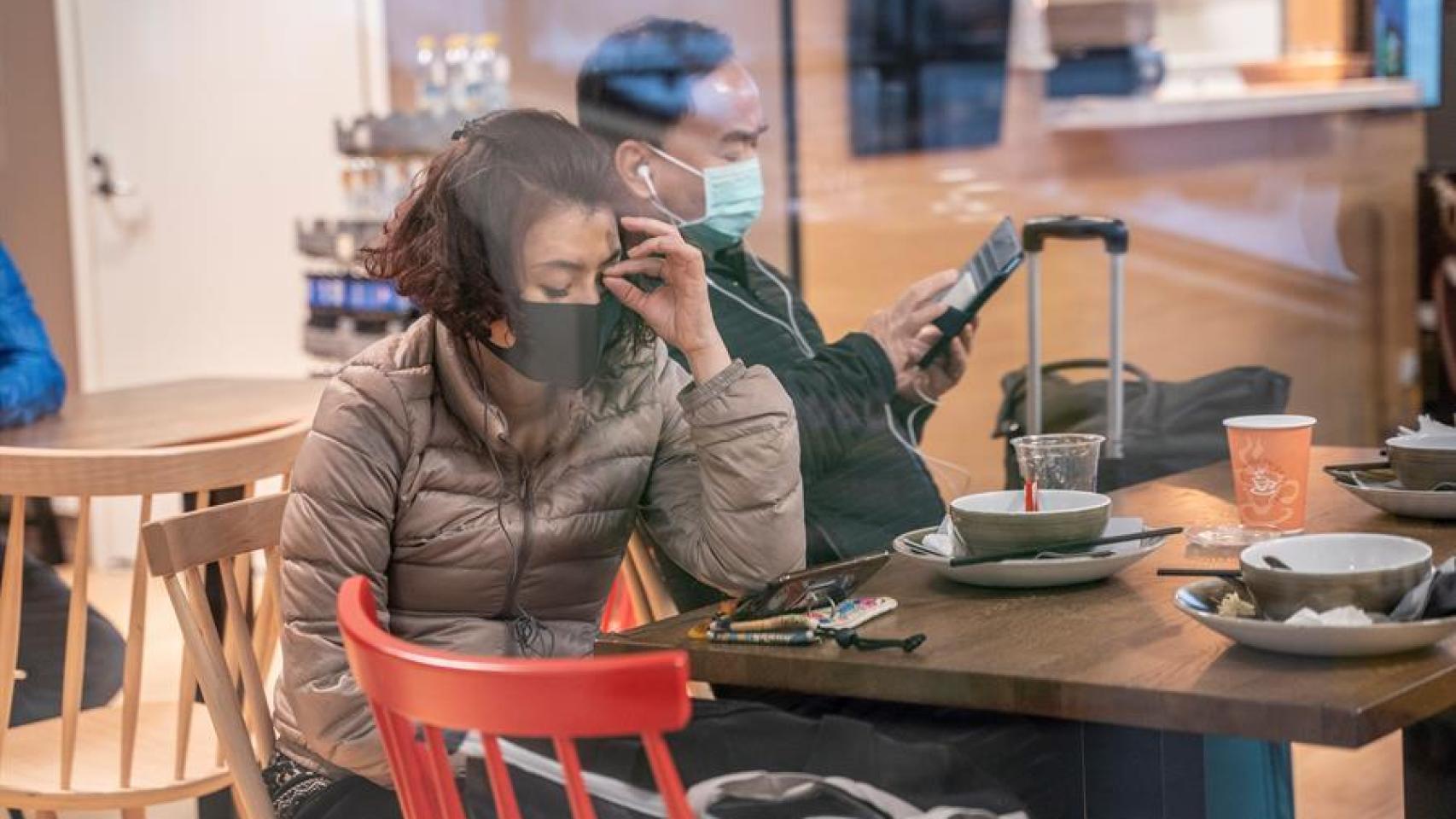 Dos personas esperan en un aeropuerto con sus mascarillas.