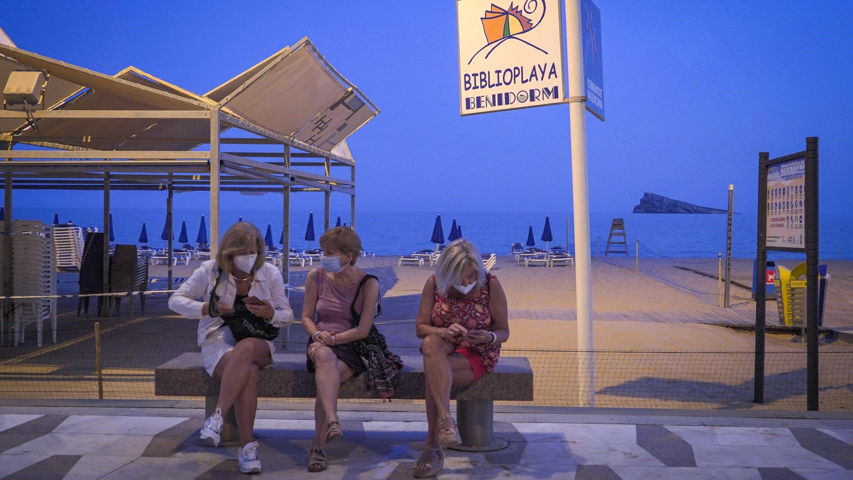Tres personas con mascarillas sentadas frente a la playa de Benidorm.