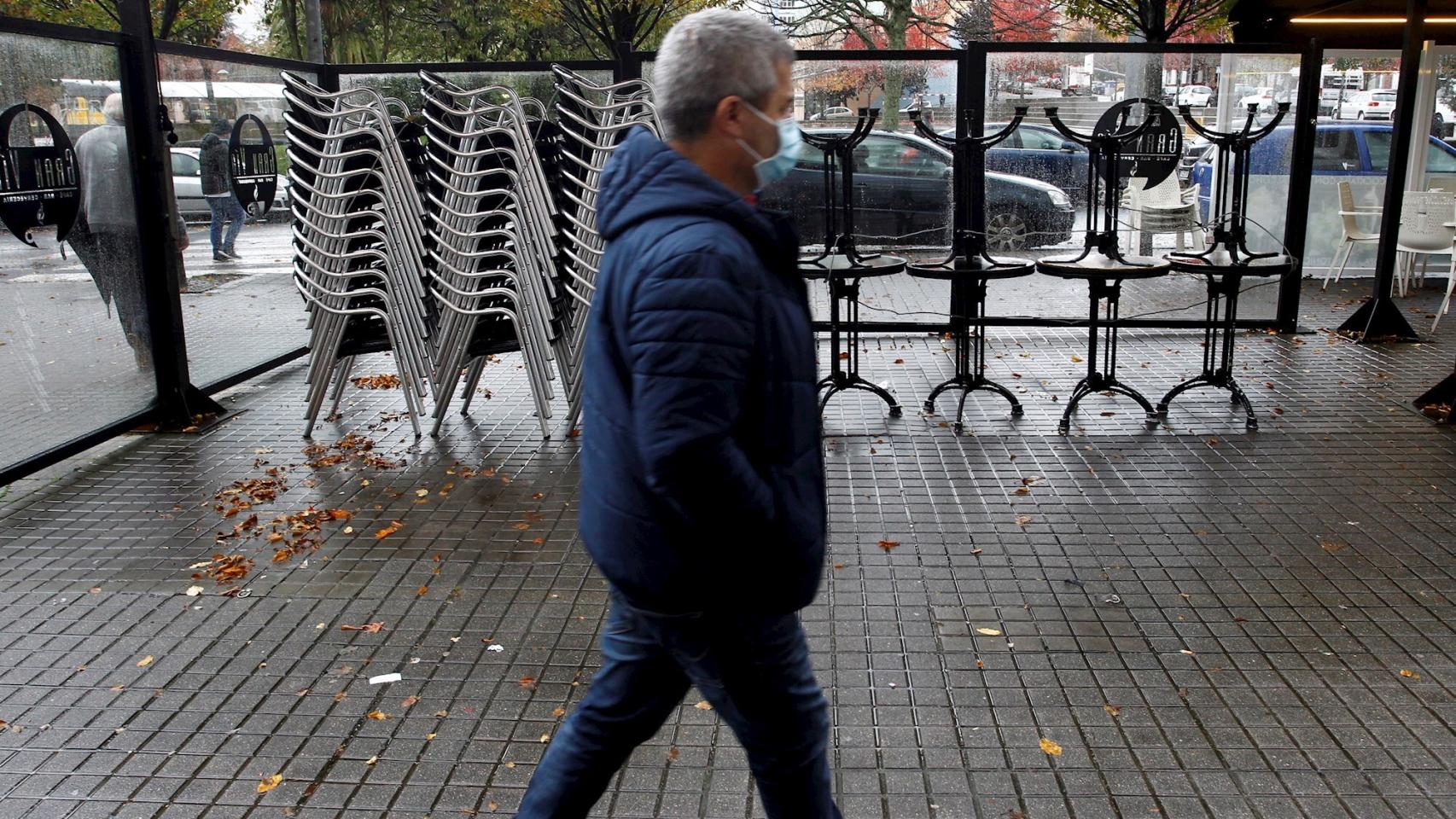 Un hombre camina por delante de las sillas y las mesas apiladas en una terraza cerrada en la localidad de Narón.