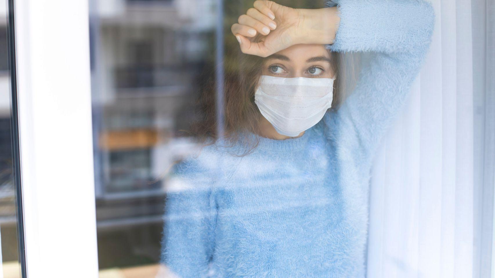 Una joven con mascarilla mirando por la ventana en actitud pensativa.