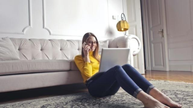 Una mujer utilizando un smartphone y un ordenador portátil sentada en el suelo de un salón.
