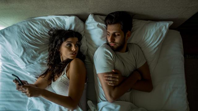 Una pareja en la cama y ella le oculta su móvil.