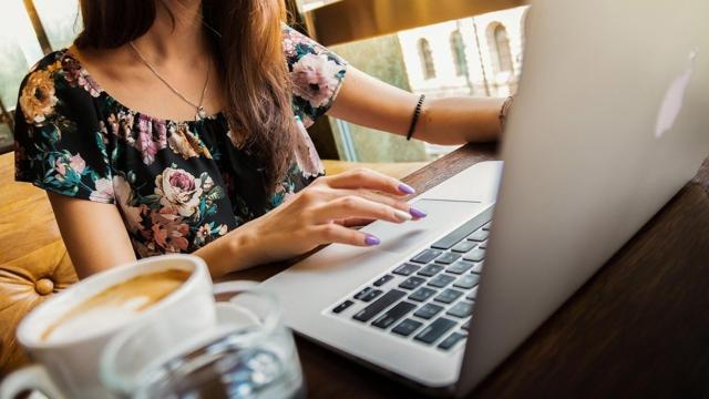 Una mujer trabajando con su ordenador junto a  una taza de café.