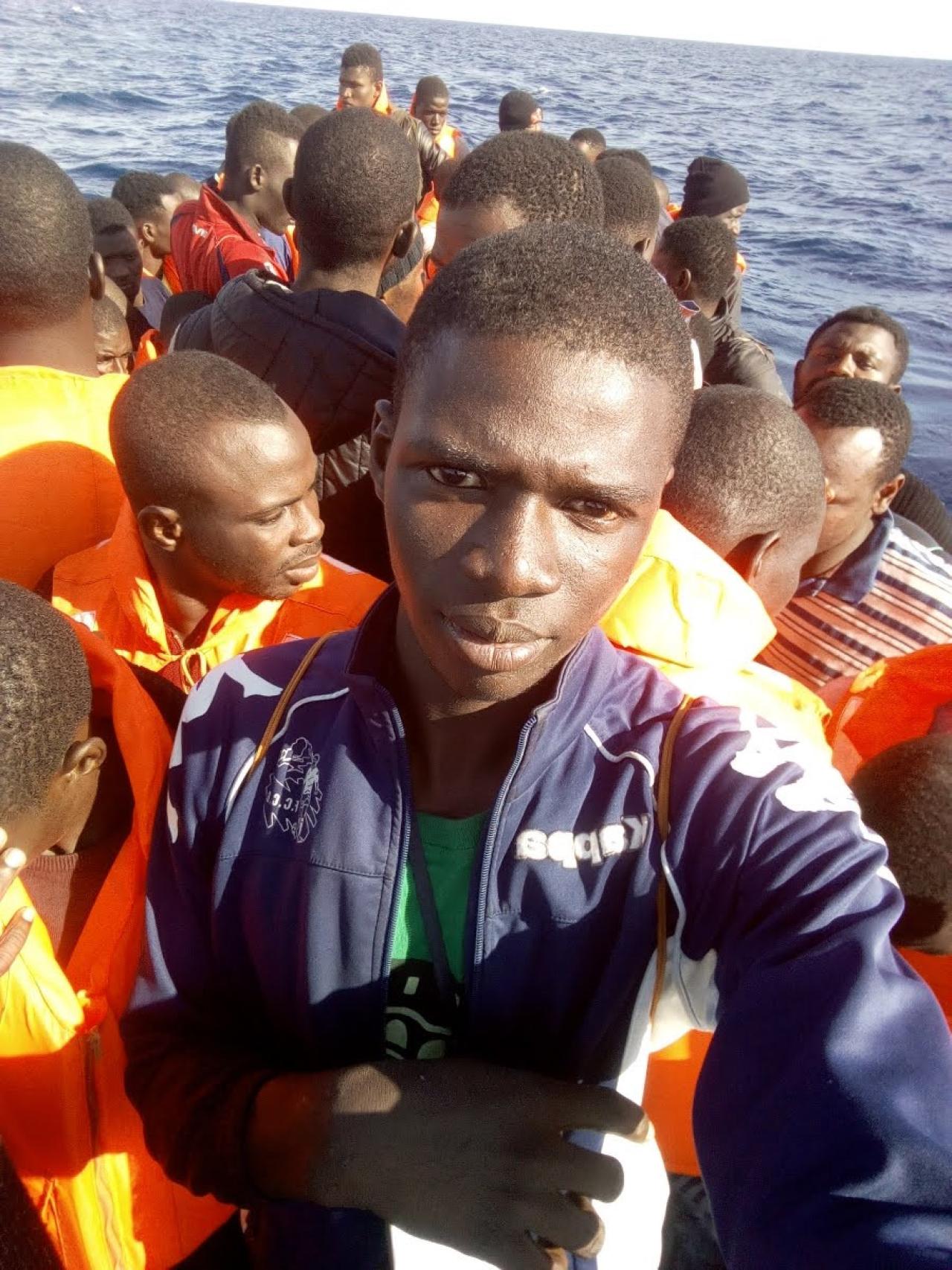 El rostro de Mory, al fondo, a la derecha del selfi que se hace el tripulante de la patera donde viajan más de veinte personas rumbo a España.