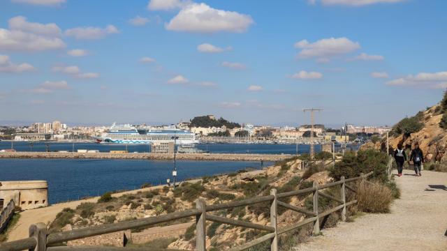 Dos senderistas caminando por el Sendero Azul Puerto de Cartagena, con un crucero atracado al fondo.