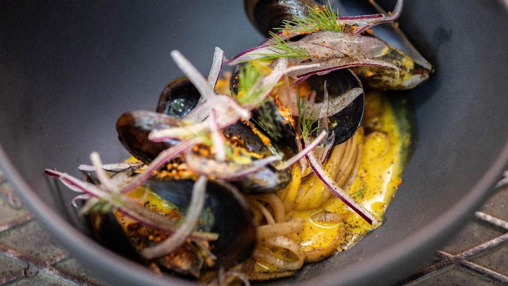 Su clásico ceviche caliente con mejillones al wok con leche de tigre de aji amarillo, noodles, migas de panko y chile, té negro lapsang souchong, cebolla roja y eneldo.