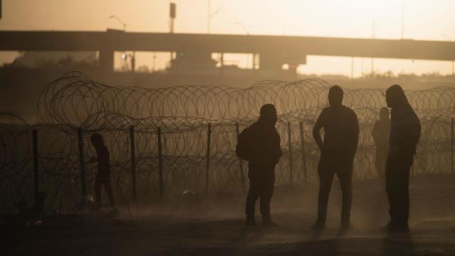 Migrantes de pie en un campo polvoriento cerca de un muro de alambre de concertina, cerca de El Paso (Texas).