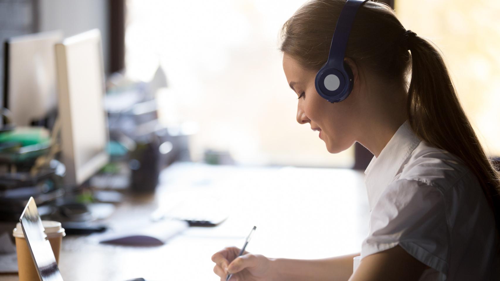 Una mujer escribe notas en su cuaderno mientras escucha a través de sus auriculares.