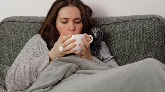 Imagen de una mujer tomando una infusión en el sofá tapada con una manta.