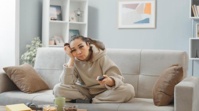 mujer-joven-ropa-casa-sentada-sofa-interior-su-casa-comiendo-galletas-sosteniendo-control-remoto-viendo-television-aburrida-triste