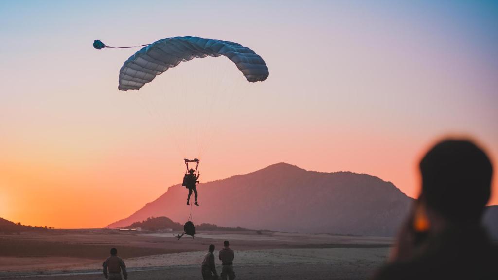 Un paracaidista del EZAPAC se prepara para tocar tierra. Se encuentra en el aire con su chaleco porta placas y fusil al tiempo que lleva la mochila de equipamiento descolgada para poder maniobrar la gran campana de los paracaídas usados.