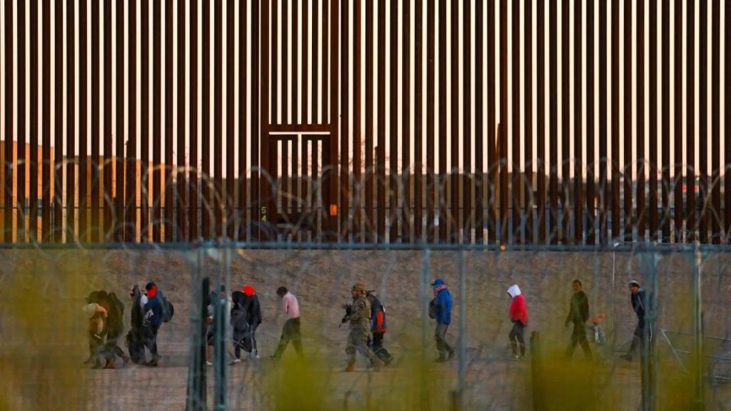 Migrantes que buscan asilo en Estados Unidos caminan cerca del muro fronterizo después de cruzar una cerca de alambre de púas desplegada para impedir su cruce hacia Estados Unidos, mientras un miembro de la Guardia Nacional de Texas los escolta.