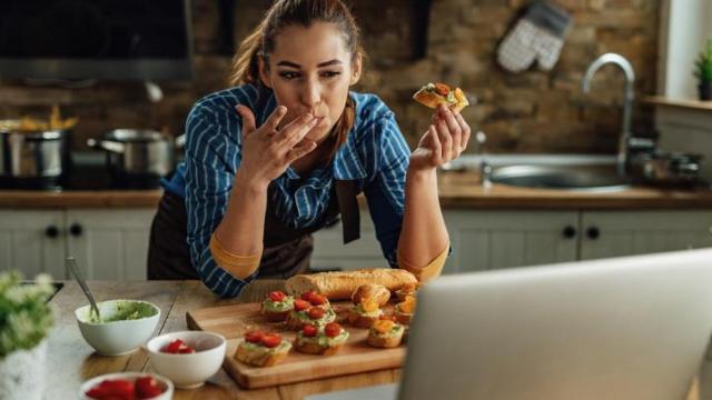 Mujer comiendo sobre la encimera de la cocina mientras mira su ordenador.