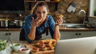 Mujer comiendo sobre la encimera de la cocina mientras mira su ordenador.