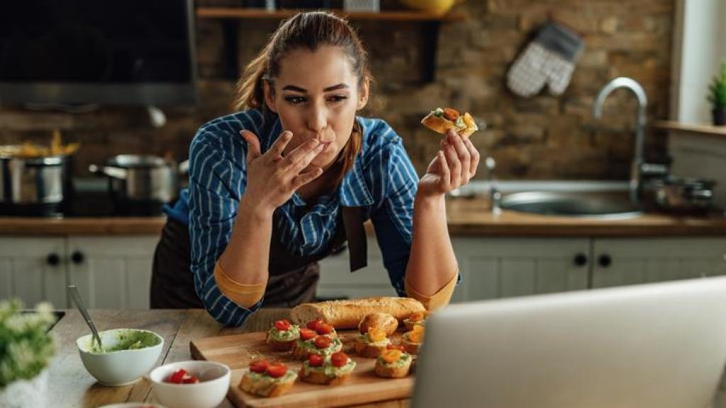 Mujer comiendo sobre la encimera de la cocina mientras mira su ordenador.