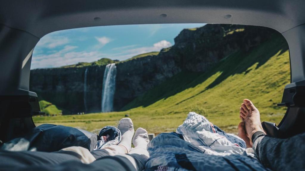 Dos personas tumbadas en la cama de su coche frente a un paisaje en la naturaleza.
