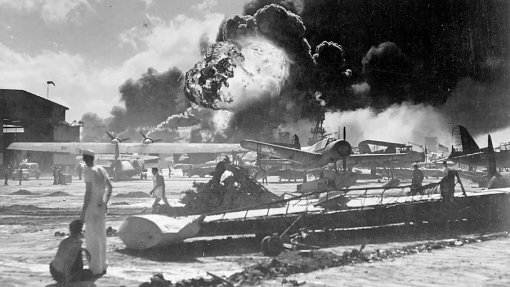 Soldados de la marina estadounidenses al lado de aviones incapacitados. Mientras, al fondo de la imagen, se aprecia la explosión de uno de los navíos.