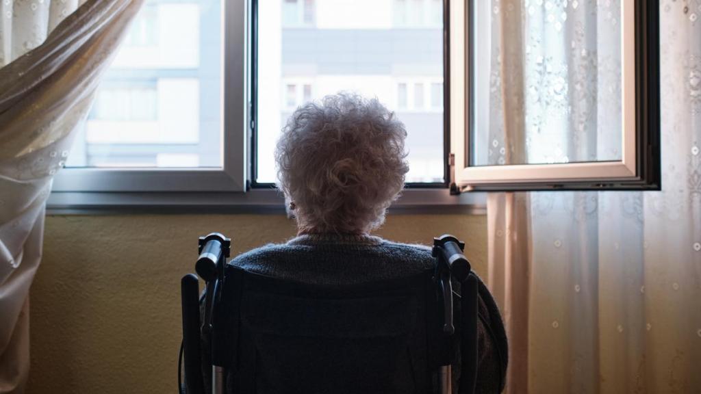 Una mujer mayor sentada mirando por la ventana de su casa.
