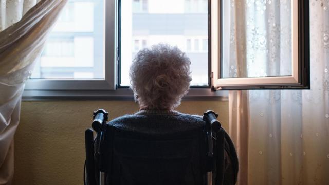 Una mujer mayor sentada mirando por la ventana de su casa.