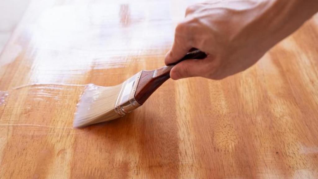 Una persona aplicando tinte transparente a una mesa de madera con pincel.