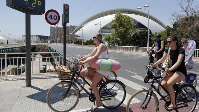 Varias jóvenes transportan sus flotadores en bici al paso de un termómetro en Valencia.