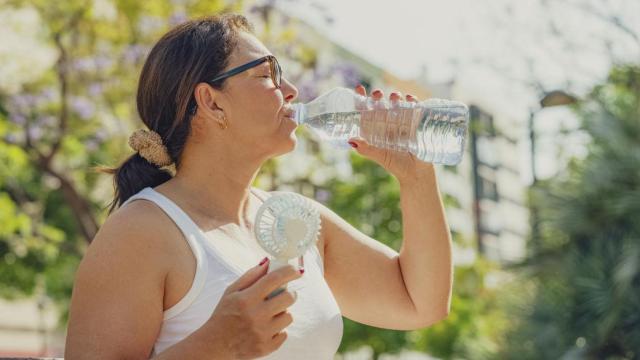 Una mujer bebe agua mientras utiliza un ventilador portátil.