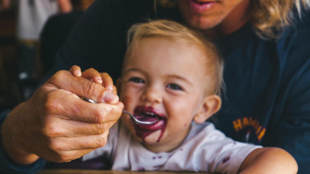 Un bebé comiendo con la ayuda de su padre.