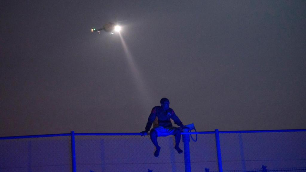 Un migrante africano se sienta en lo alto de una valla fronteriza cubierta de alambre de espino entre Marruecos y Melilla.