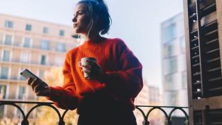 Mujer joven en el balcón con el móvil y un café.