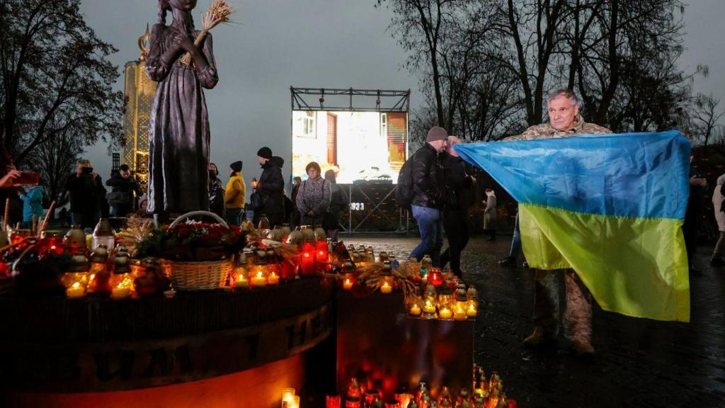 Un hombre sostiene una bandera ucraniana mientras la gente coloca velas, flores y gavillas de trigo en el monumento Amargo Recuerdo de la Infancia con la estatua de la Niña con Espigas en el complejo del Museo Nacional del Genocidio del Holodomor en Kiev.