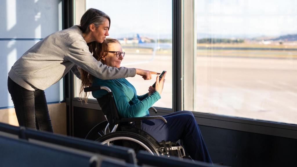 Mujer asistiendo a un usuario de silla de ruedas con un teléfono inteligente en el aeropuerto.