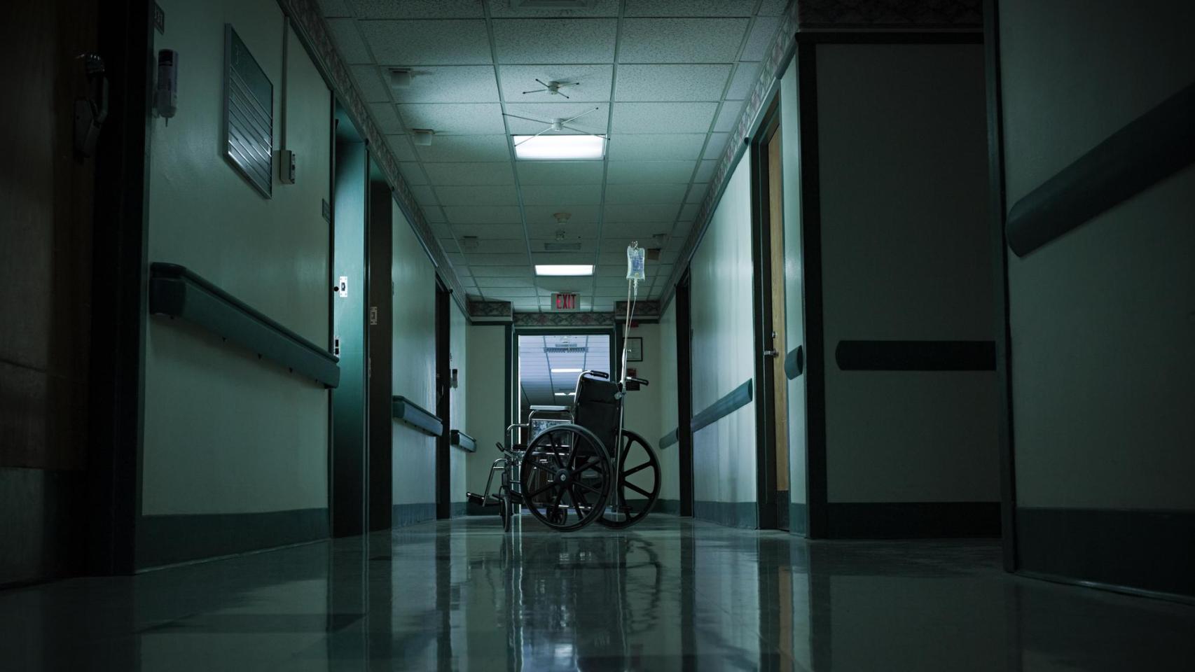Pasillo oscuro de un hospital con una silla de ruedas al fondo.