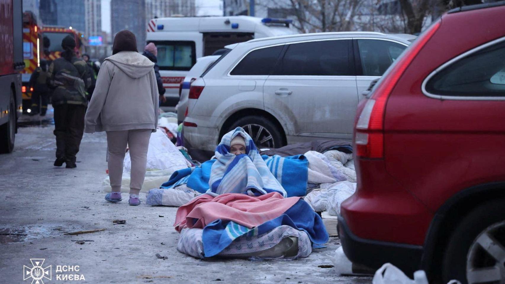 Una mujer sentada en el suelo cubierta con mantas frente a un edificio destruido por un ataque ruso.