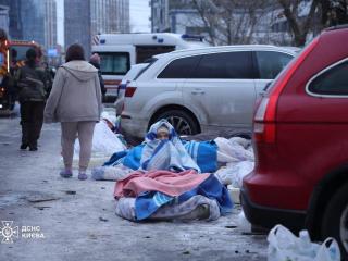 A woman sits on the ground covered in blankets in front of a building destroyed by a Russian attack.