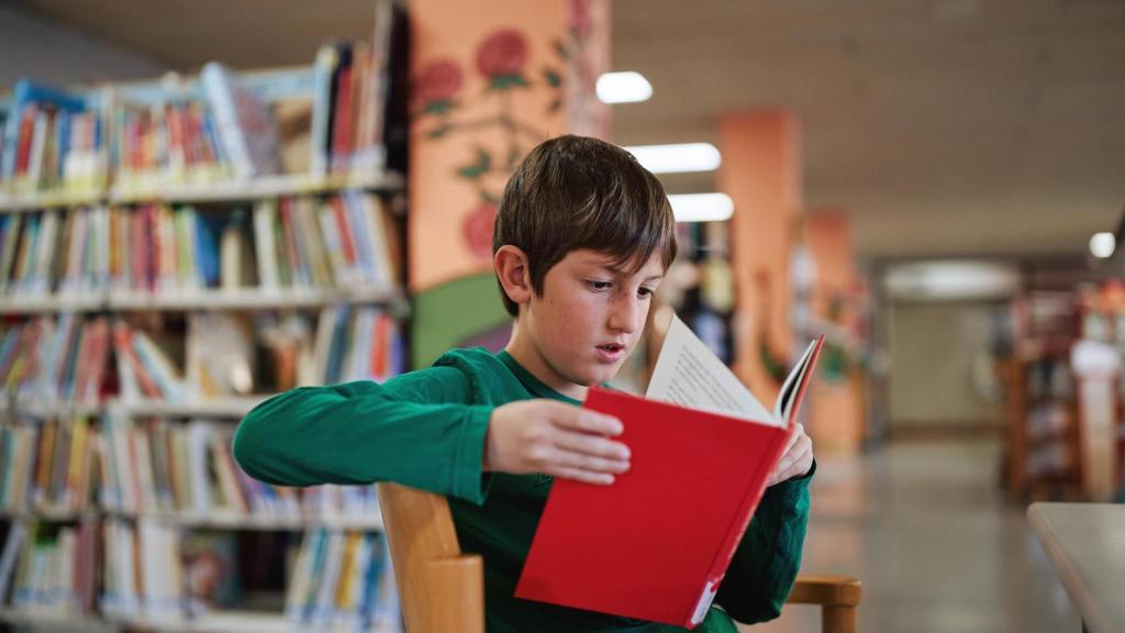 Niño leyendo un libro rojo en una biblioteca.