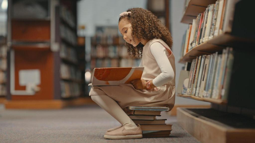 Niña sentada en libros en la biblioteca y leyendo un libro.