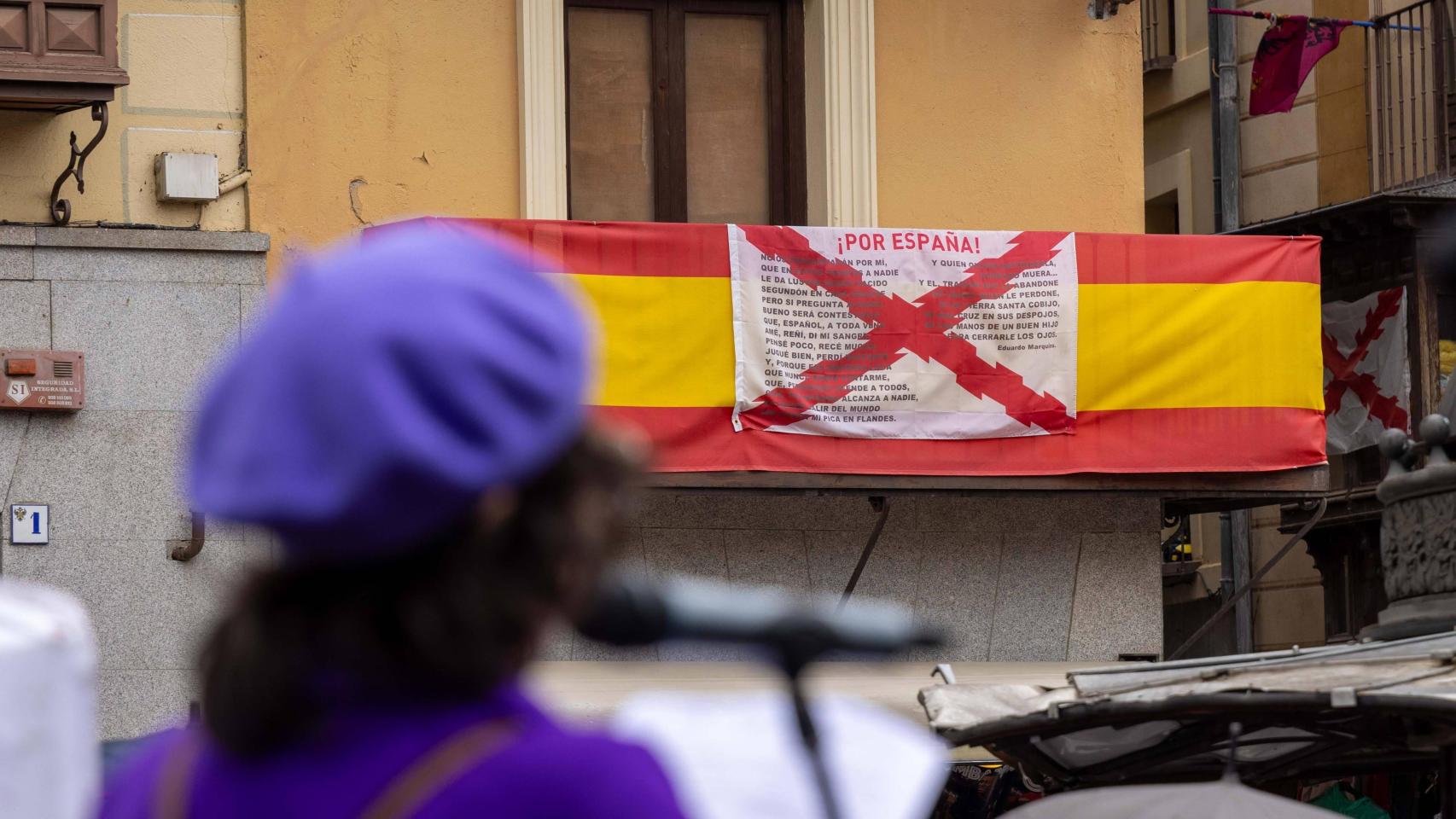 Una manifestante en la marcha del 8M de 2025 junto a una balconada de Toledo en la que cuelgan unas banderas de España y de la Cruz de Borgoña.