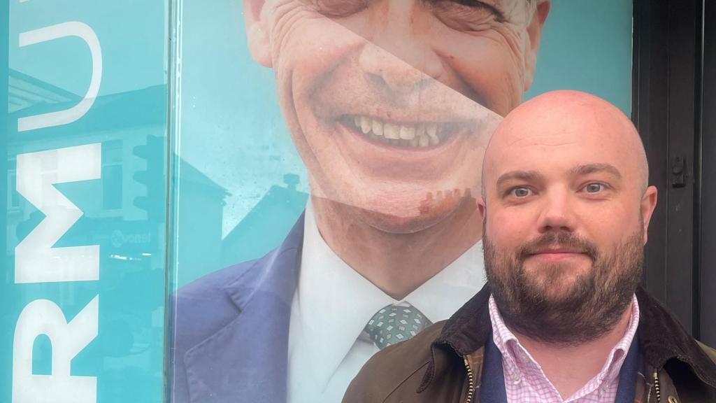 El candidato de Welsh Reform UK, Llyr Powell, posa frente a una foto del líder del partido, Nigel Farage.