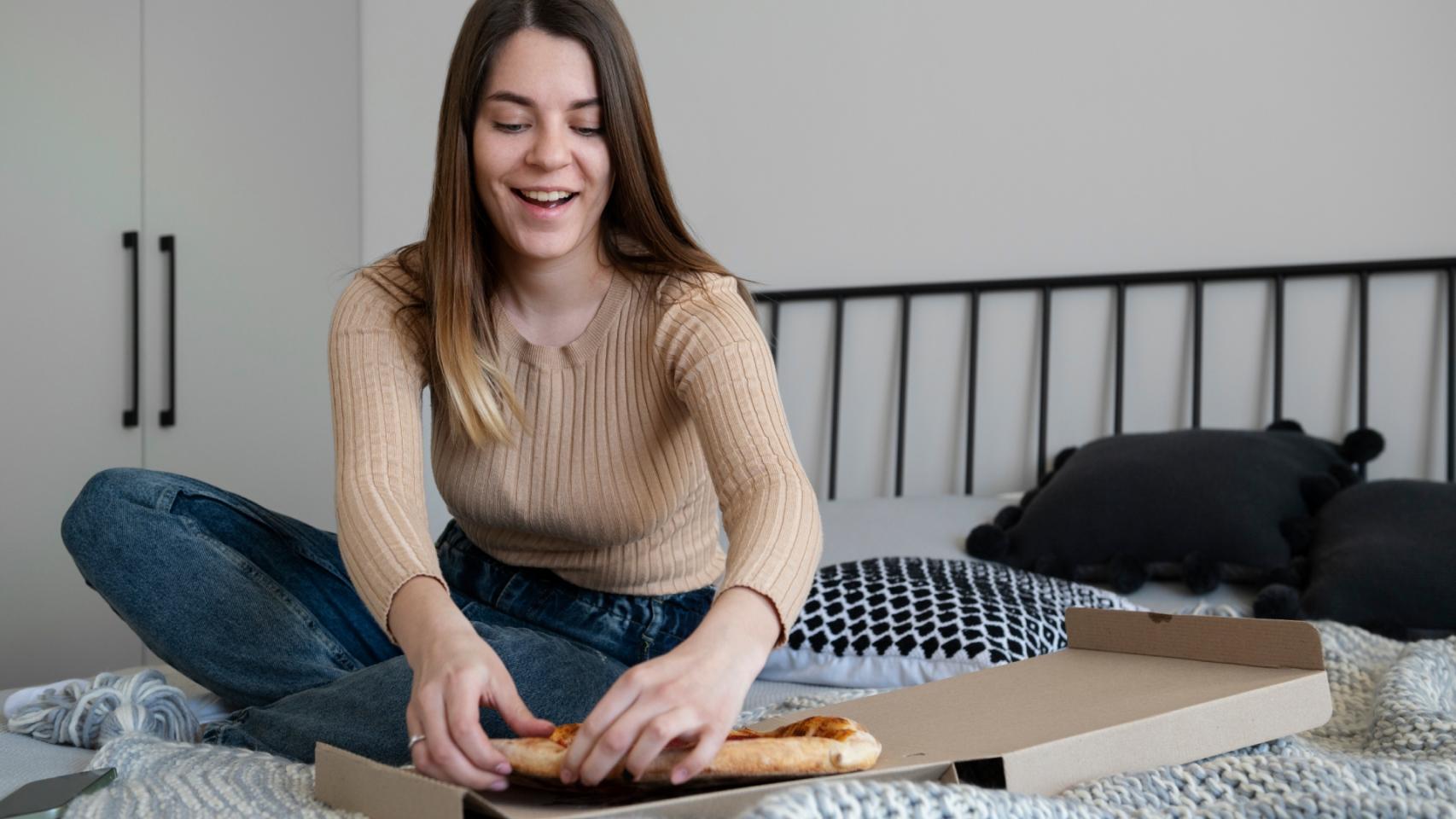 Una joven se come un trozo de pizza en su habitación