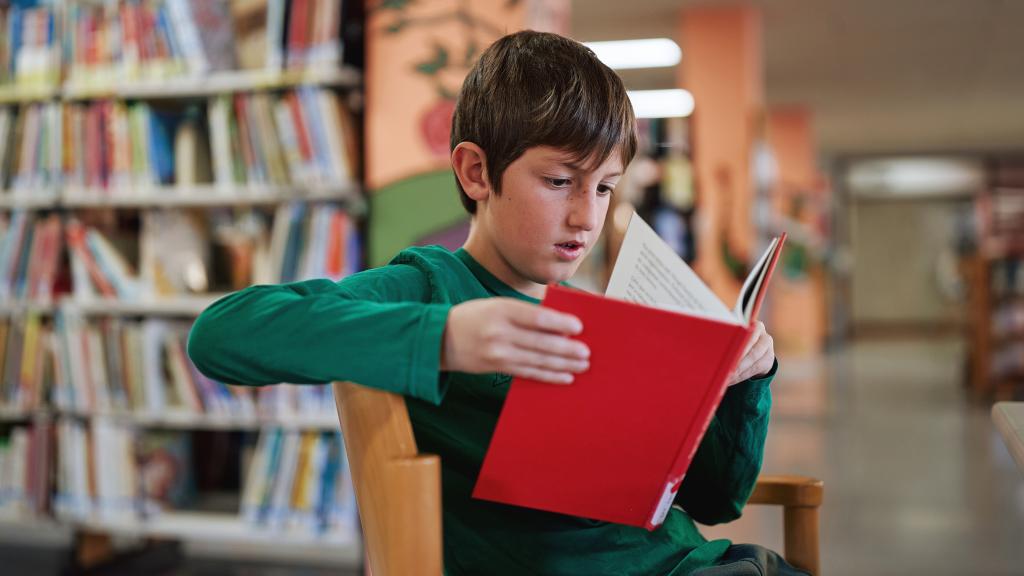 Niño leyendo un libro rojo en una biblioteca.