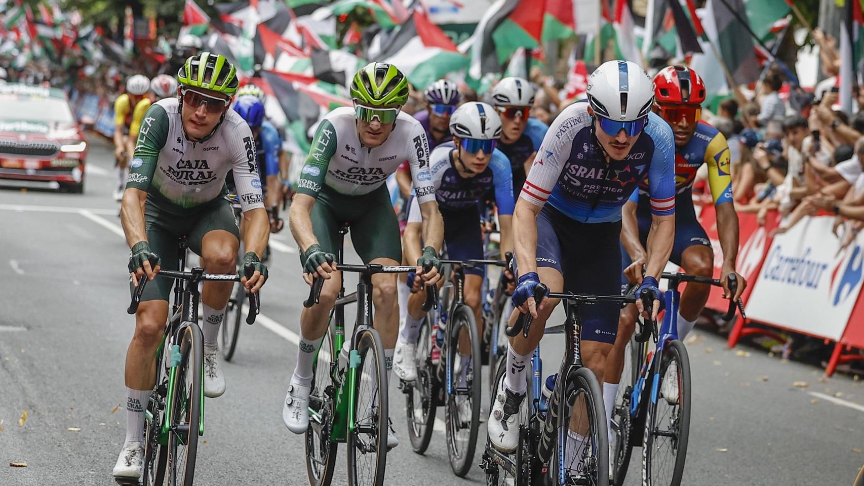 Un miembro del Israel Premier Tech en La Vuelta 2025 y, al fondo, una protesta propalestina